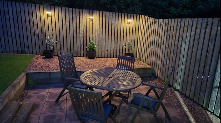 Paved patio with a round wooden table and chairs, timber fencing and wall lights at dusk.