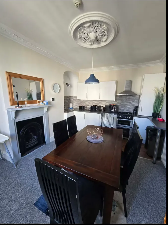 Kitchen with white cabinets, a dining table and chairs, fireplace and ornate ceiling rose.