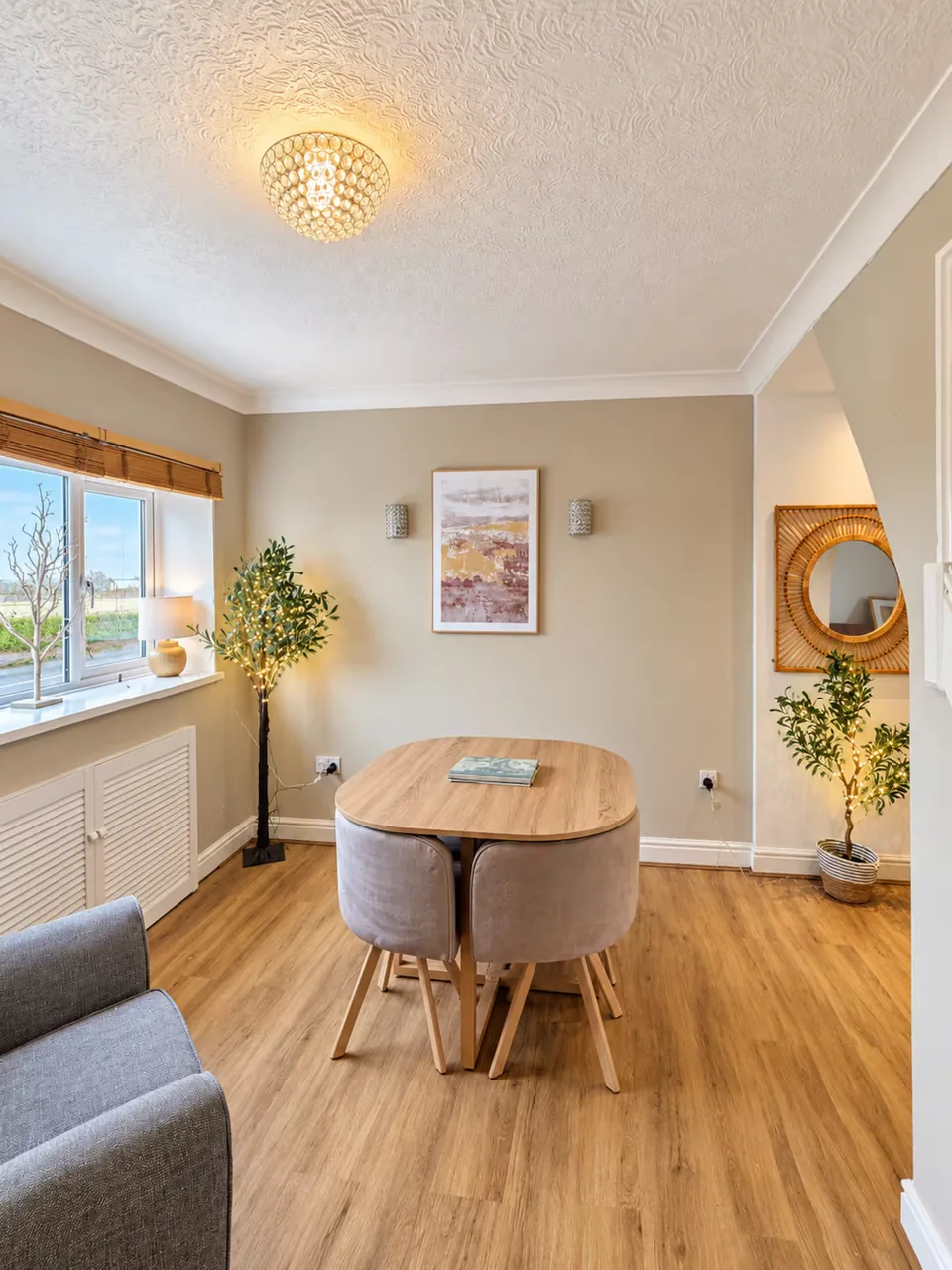 Wooden dining table and upholstered chairs beside a window, with soft lighting and decorative trees.
