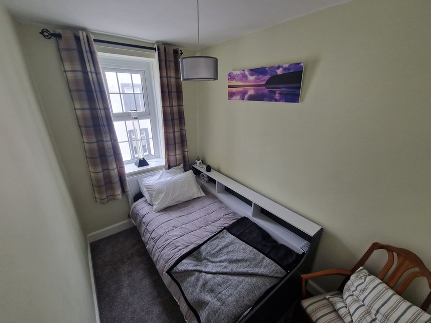 Single bed beside a window with a bubble tube on the sill, low shelving and a cushioned wooden chair.