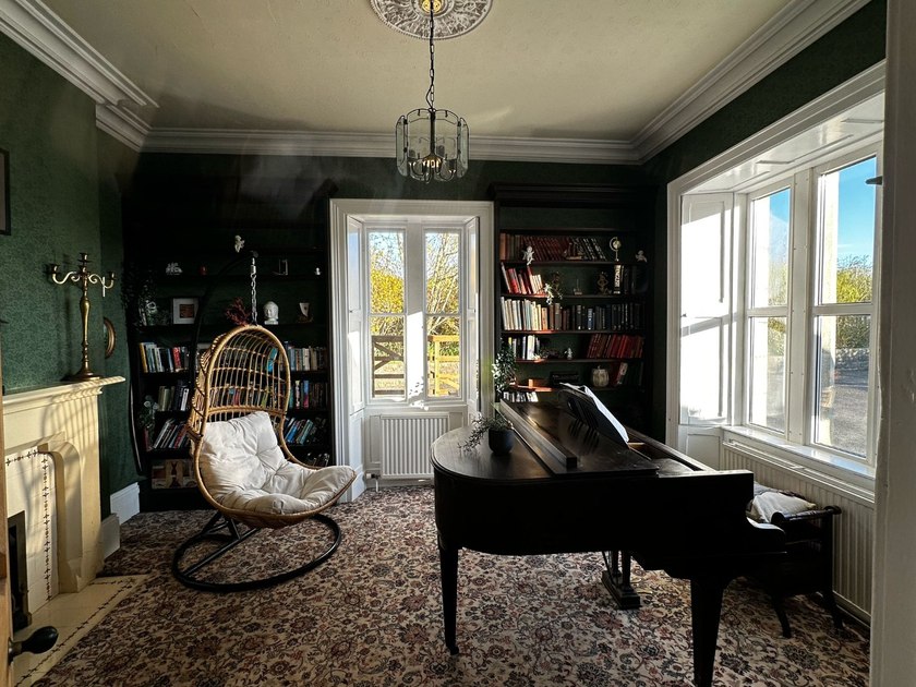 A dark wooden grand piano, a hanging wicker chair and bookcases in a green-walled room with two windows and a fireplace.