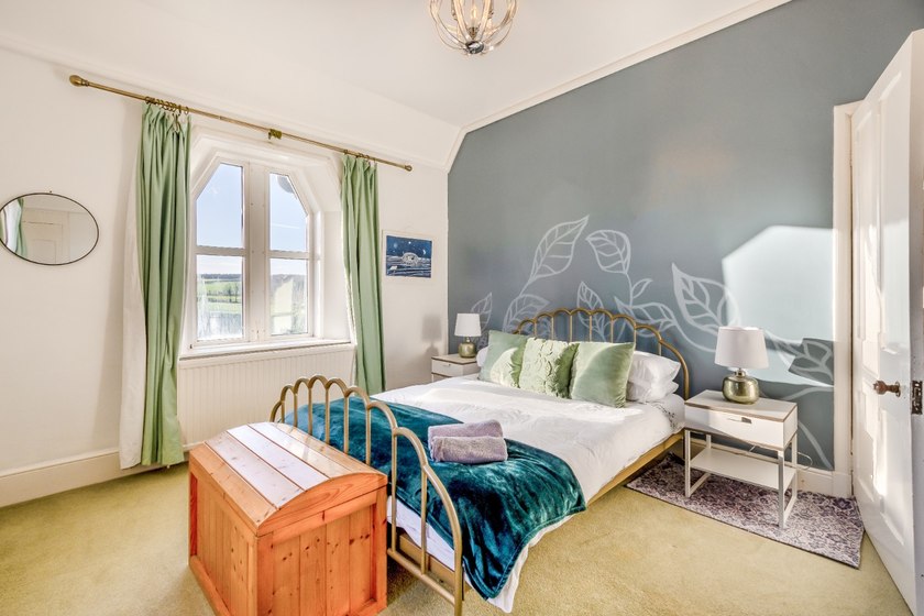 A brass-coloured bed with a teal throw and green cushions stands against a blue-grey leaf-patterned wall, beside a window with green curtains.